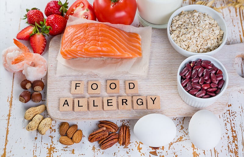 A variety of common food allergens, including fish, eggs, nuts, dairy, and strawberries, arranged around wooden tiles spelling out "FOOD ALLERGY," emphasising awareness and safety regarding dietary restrictions and food sensitivities.