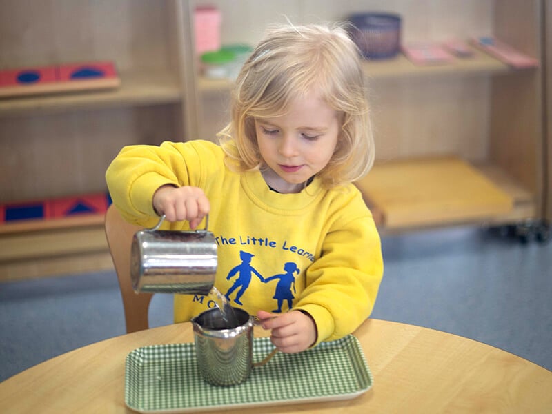 A young child at The Little Learners Montessori engaged in a practical life activity, carefully pouring water from one jug to another, demonstrating the Montessori practical life ethos of fostering independence and fine motor skills.