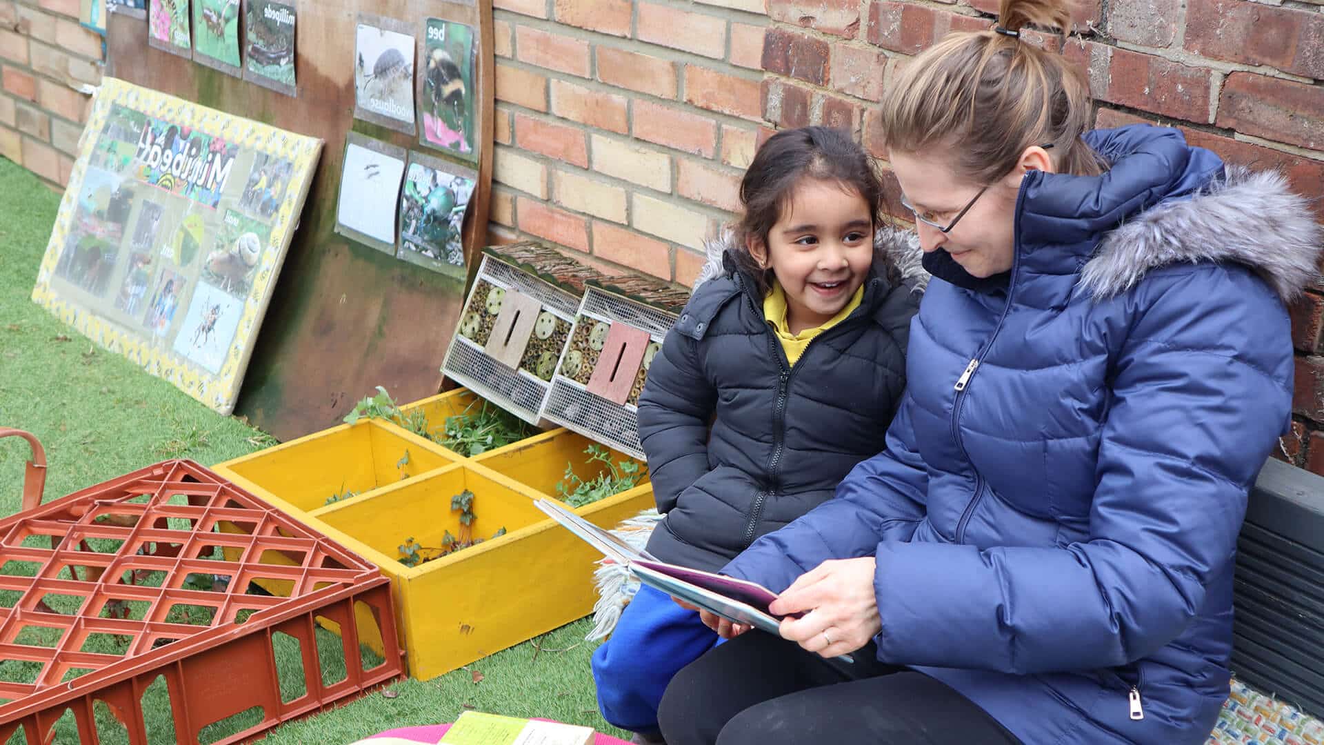 A child and adult sit outdoors, sharing a book, surrounded by nature-themed educational displays and insect pictures.